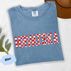 Może przedstawiać: Jasnoniebieska koszulka z napisem "HRVATSKA" w czerwono-białą kratę. Koszulka ma okrągły dekolt i metkę Comfort Colors. Brązowy kapelusz jest widoczny w prawym g&oacute;rnym rogu.