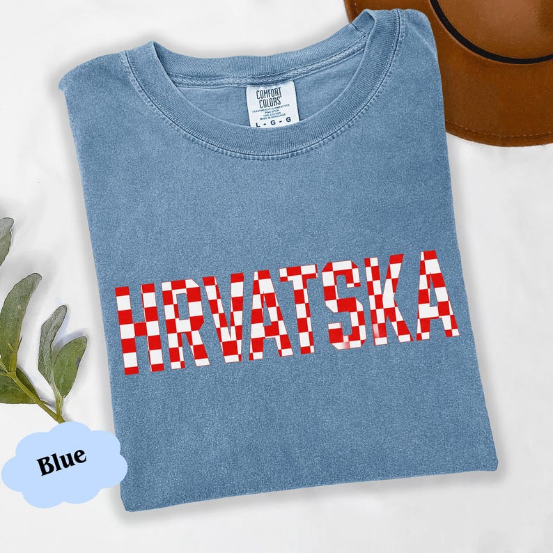 Może przedstawiać: Jasnoniebieska koszulka z napisem "HRVATSKA" w czerwono-białą kratę. Koszulka ma okrągły dekolt i metkę Comfort Colors. Brązowy kapelusz jest widoczny w prawym g&oacute;rnym rogu.
