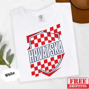 Pu&ograve; includere: Una t-shirt bianca con un design grafico che presenta un motivo a scacchi rosso e bianco all'interno del contorno di uno scudo. La parola HRVATSKA &egrave; stampata in lettere maiuscole blu scuro al centro dello scudo. La maglia ha un girocollo.