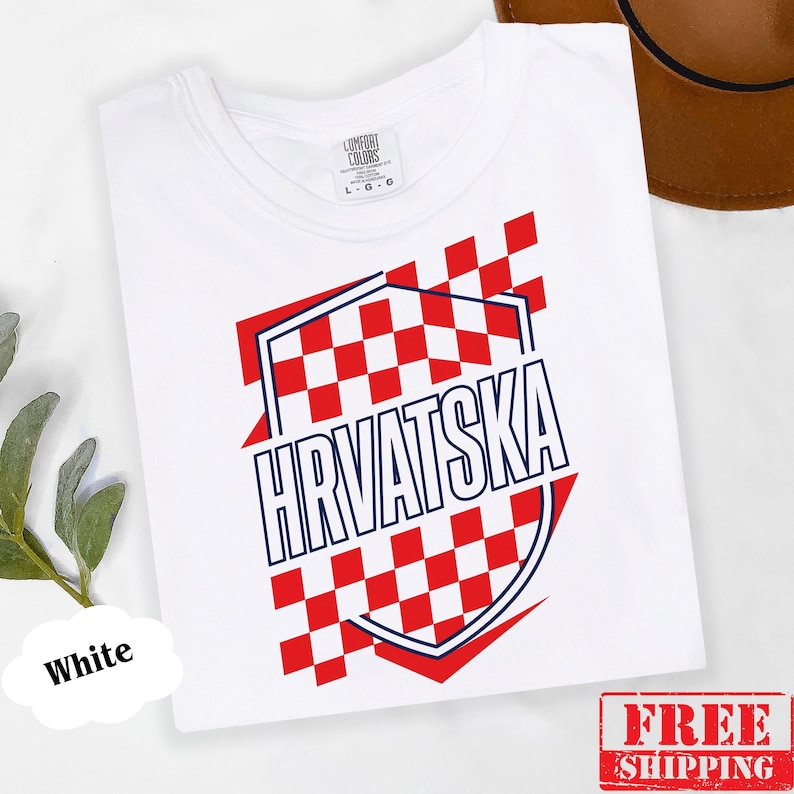 Pu&ograve; includere: Una t-shirt bianca con un design grafico che presenta un motivo a scacchi rosso e bianco all'interno del contorno di uno scudo. La parola HRVATSKA &egrave; stampata in lettere maiuscole blu scuro al centro dello scudo. La maglia ha un girocollo.