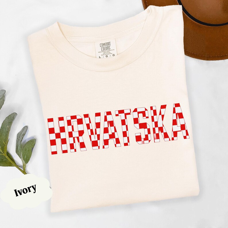 Może przedstawiać: Kremowa koszulka z napisem "HRVATSKA" w czerwono-białą szachownicę. Koszulka ma okrągły dekolt i metkę "Comfort Colors". Brązowy kapelusz widoczny w tle.
