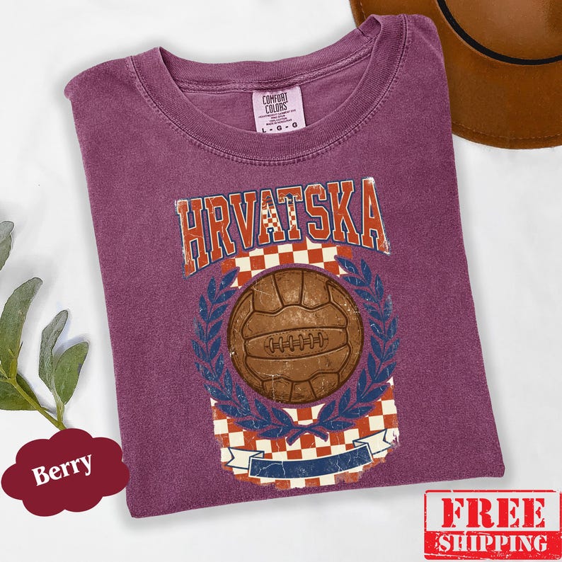 May include: A berry-colored t-shirt featuring a vintage-style graphic with the text HRVATSKA in a distressed font. The design includes a brown soccer ball centered over a red and white checkered pattern, framed by blue laurel leaves and a banner.
