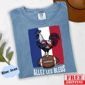 Puede incluir: Una camiseta azul de cuello redondo con un gr&aacute;fico de un gallo de pie sobre un bal&oacute;n de f&uacute;tbol americano marr&oacute;n sobre el fondo de la bandera francesa. El texto "ALLEZ LES BLEUS" est&aacute; impreso en blanco debajo del gr&aacute;fico. Etiqueta Comfort Colors.