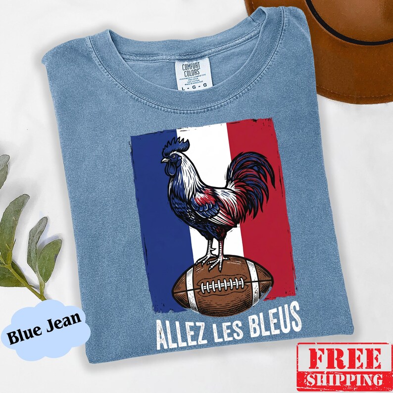 Puede incluir: Una camiseta azul de cuello redondo con un gr&aacute;fico de un gallo de pie sobre un bal&oacute;n de f&uacute;tbol americano marr&oacute;n sobre el fondo de la bandera francesa. El texto "ALLEZ LES BLEUS" est&aacute; impreso en blanco debajo del gr&aacute;fico. Etiqueta Comfort Colors.