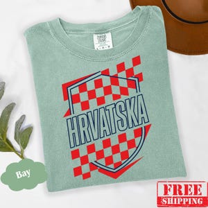 Pu&ograve; includere: Una t-shirt verde salvia con una grafica a scacchi rossi e bianchi con un contorno blu scuro. Il testo "HRVATSKA" &egrave; stampato in lettere maiuscole blu scuro al centro. La t-shirt presenta un'etichetta del marchio Comfort Colors.
