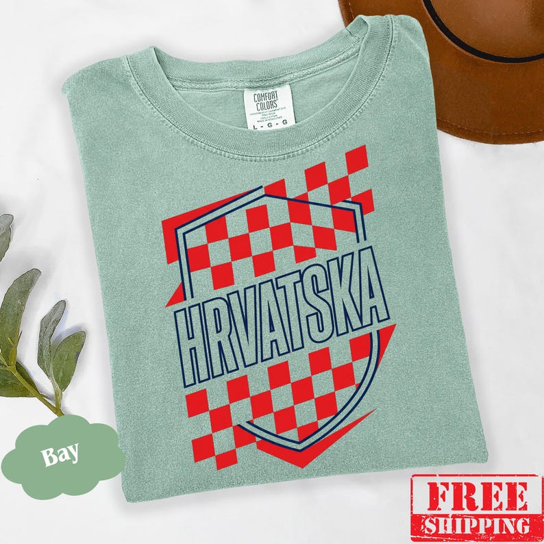 Pu&ograve; includere: Una t-shirt verde salvia con una grafica a scacchi rossi e bianchi con un contorno blu scuro. Il testo "HRVATSKA" &egrave; stampato in lettere maiuscole blu scuro al centro. La t-shirt presenta un'etichetta del marchio Comfort Colors.