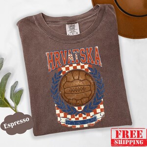 May include: An espresso-colored t-shirt featuring a vintage-style graphic with the text HRVATSKA in red and white. The design includes a brown soccer ball centered over a red and white checkered pattern, framed by blue laurel leaves and a banner.