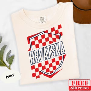 Pu&ograve; includere: Una t-shirt a maniche corte color avorio con un motivo a scacchi rossi e bianchi a forma di scudo. La parola HRVATSKA &egrave; stampata in lettere maiuscole blu scuro al centro del disegno. La maglietta presenta un classico girocollo.