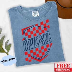 Pu&ograve; includere: Una t-shirt color blu jeans con un design grafico a scacchi rossi e bianchi all'interno di una forma a scudo con contorno blu scuro. La parola HRVATSKA &egrave; stampata in grandi lettere maiuscole blu scuro al centro dello scudo.