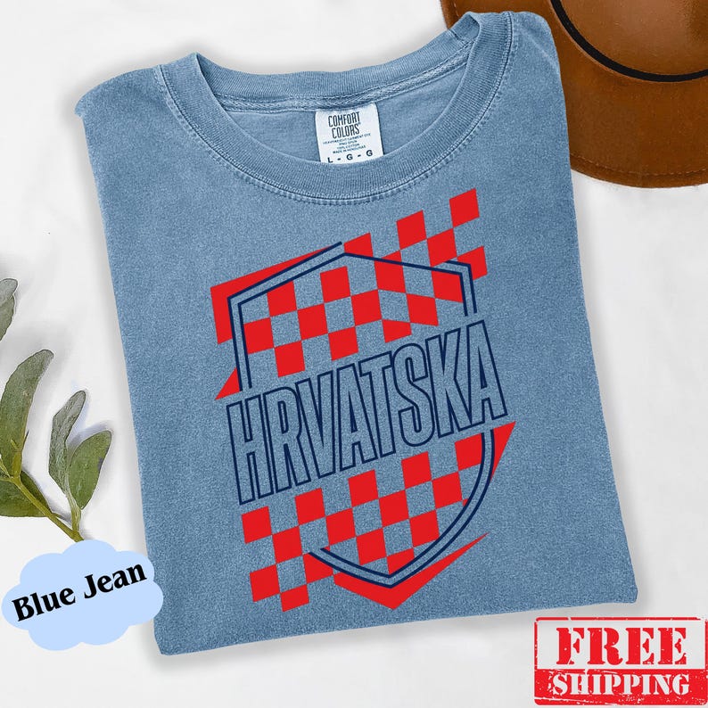 Pu&ograve; includere: Una t-shirt color blu jeans con un design grafico a scacchi rossi e bianchi all'interno di una forma a scudo con contorno blu scuro. La parola HRVATSKA &egrave; stampata in grandi lettere maiuscole blu scuro al centro dello scudo.