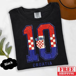 Puede incluir: Una camiseta negra de manga corta con un gran gr&aacute;fico desgastado del n&uacute;mero 10 relleno con el patr&oacute;n de la bandera croata roja, blanca y azul y su escudo de armas. La palabra CROATIA est&aacute; impresa en azul debajo del n&uacute;mero. Etiqueta Comfort Colors.