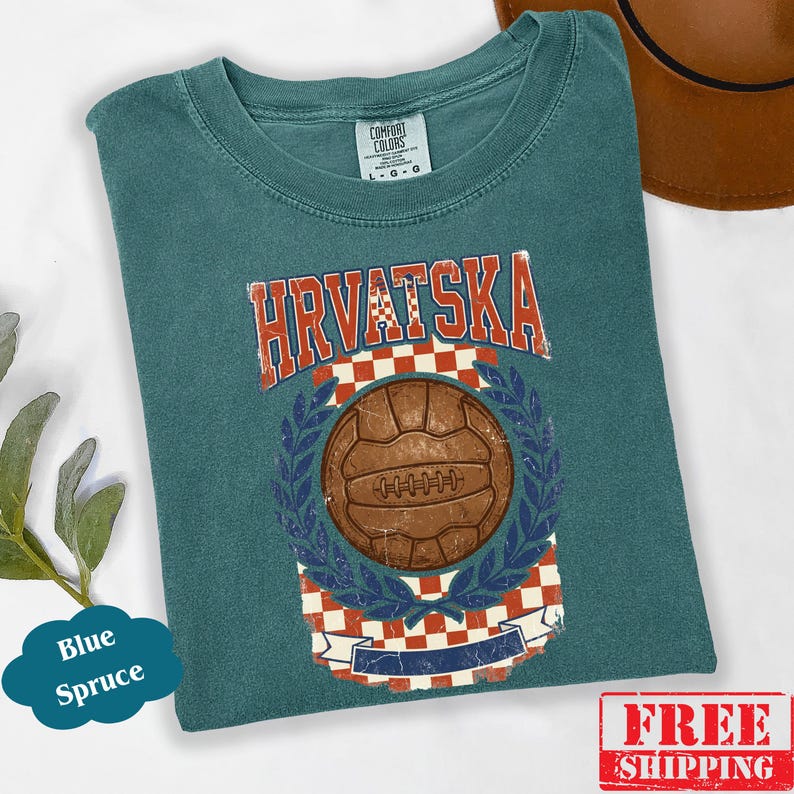 May include: A blue spruce t-shirt featuring a vintage-style graphic with the text HRVATSKA in red and white. The design includes a brown soccer ball centered within a red and white checkered pattern, framed by blue laurel leaves and a banner.