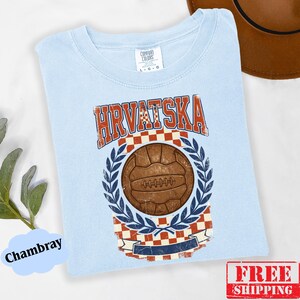 May include: A light blue t-shirt featuring a vintage-style graphic with the text HRVATSKA in red, a brown leather soccer ball, a red and white checkered pattern, and blue laurel leaves. The shirt has a crew neckline and a Comfort Colors brand label.