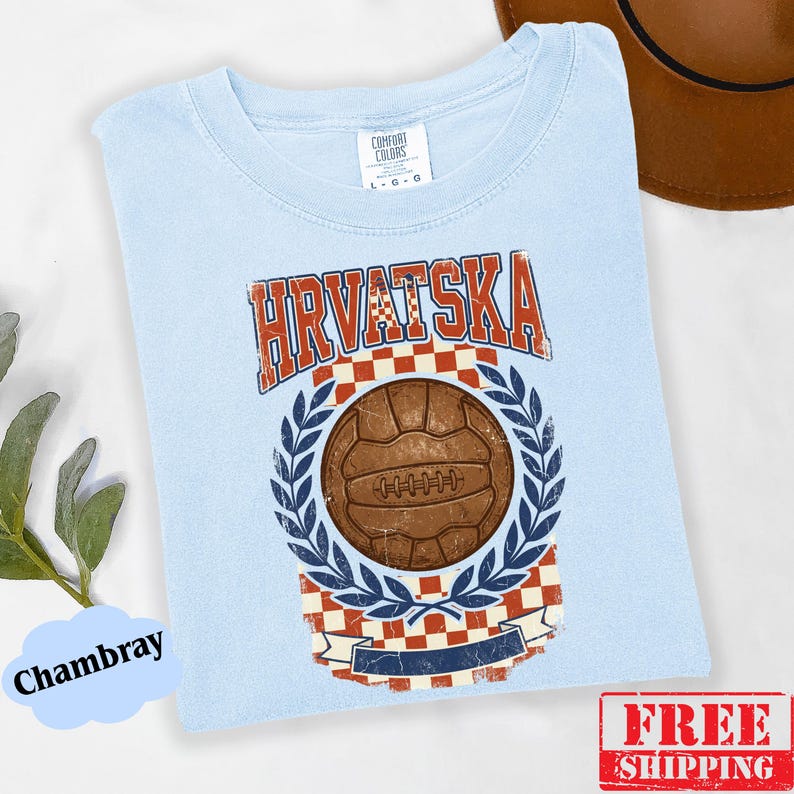 May include: A light blue t-shirt featuring a vintage-style graphic with the text HRVATSKA in red, a brown leather soccer ball, a red and white checkered pattern, and blue laurel leaves. The shirt has a crew neckline and a Comfort Colors brand label.
