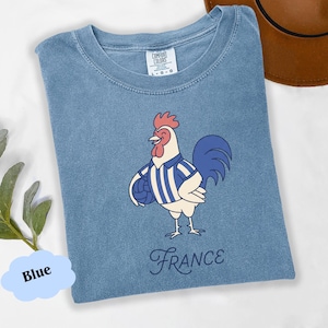 May include: A blue t-shirt with a cartoon rooster wearing a striped shirt and holding a ball. The word "France" is written in a script font below the rooster. The shirt has a crew neck and short sleeves.