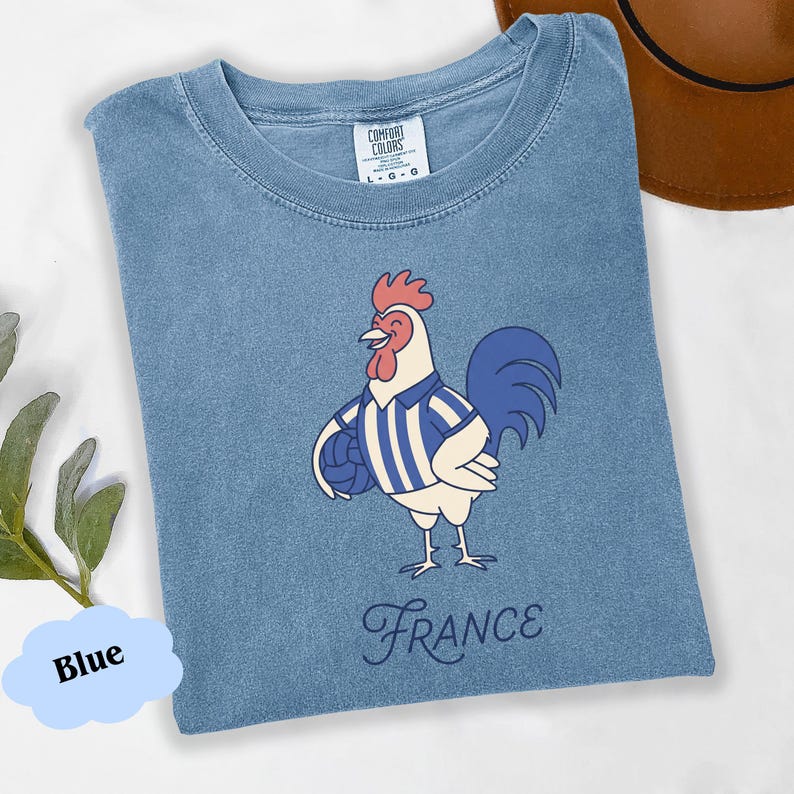 May include: A blue t-shirt with a cartoon rooster wearing a striped shirt and holding a ball. The word "France" is written in a script font below the rooster. The shirt has a crew neck and short sleeves.