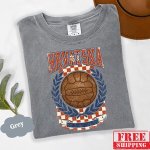 May include: A grey short-sleeve t-shirt featuring a vintage-style graphic with the text HRVATSKA in red and white. The design includes a brown soccer ball centered within a blue laurel wreath, set against a red and white checkered background.