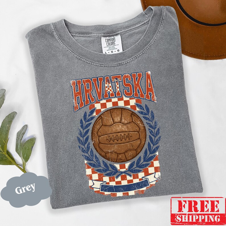 May include: A grey short-sleeve t-shirt featuring a vintage-style graphic with the text HRVATSKA in red and white. The design includes a brown soccer ball centered within a blue laurel wreath, set against a red and white checkered background.