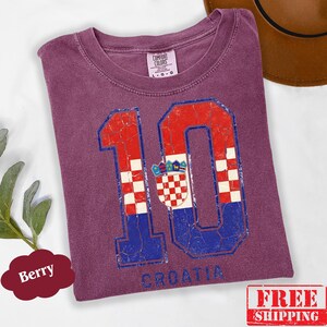 Puede incluir: Una camiseta color baya con un gr&aacute;fico grande y desgastado del n&uacute;mero 10. El n&uacute;mero est&aacute; relleno con un patr&oacute;n de cuadros rojos y blancos y secciones azules, incorporando el escudo de armas croata. La palabra CROATIA est&aacute; impresa debajo.