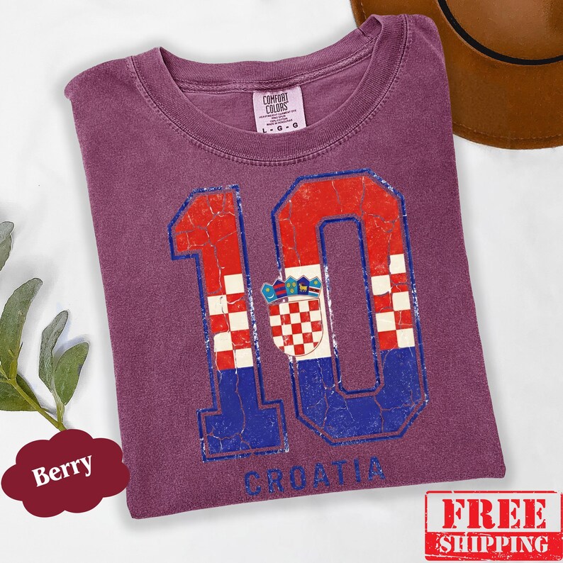 Puede incluir: Una camiseta color baya con un gr&aacute;fico grande y desgastado del n&uacute;mero 10. El n&uacute;mero est&aacute; relleno con un patr&oacute;n de cuadros rojos y blancos y secciones azules, incorporando el escudo de armas croata. La palabra CROATIA est&aacute; impresa debajo.