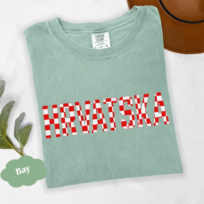 Może przedstawiać: Jasnozielona koszulka z napisem "HRVATSKA" w czerwono-białą kratę. Koszulka marki Comfort Colors, rozmiar L-G-G. Brązowy kapelusz widoczny w prawym g&oacute;rnym rogu.