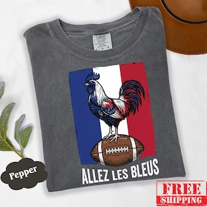 Puede incluir: Una camiseta gris carb&oacute;n con un gr&aacute;fico de un gallo de pie sobre un bal&oacute;n de f&uacute;tbol americano frente a un fondo de bandera azul, blanca y roja. El texto "ALLEZ LES BLEUS" est&aacute; impreso en blanco debajo del gr&aacute;fico. Etiqueta Comfort Colors.