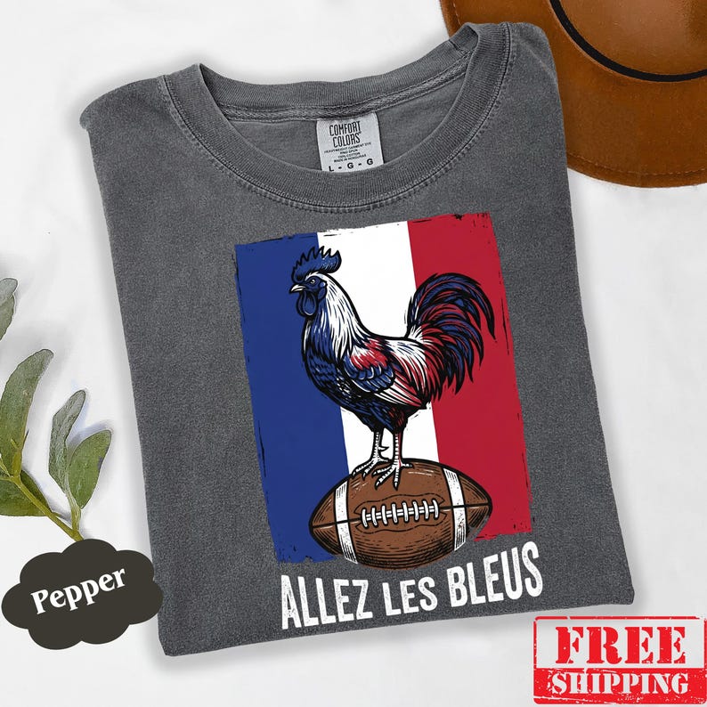 Puede incluir: Una camiseta gris carb&oacute;n con un gr&aacute;fico de un gallo de pie sobre un bal&oacute;n de f&uacute;tbol americano frente a un fondo de bandera azul, blanca y roja. El texto "ALLEZ LES BLEUS" est&aacute; impreso en blanco debajo del gr&aacute;fico. Etiqueta Comfort Colors.