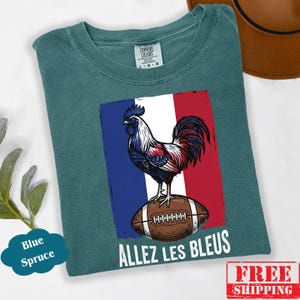 Puede incluir: Una camiseta de color azul verdoso con un gr&aacute;fico de un gallo de pie sobre un bal&oacute;n de f&uacute;tbol americano marr&oacute;n sobre el fondo de la bandera francesa. El texto "ALLEZ LES BLEUS" est&aacute; impreso en blanco debajo del bal&oacute;n. Tiene una etiqueta Comfort Colors.