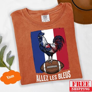 Puede incluir: Una camiseta de manga corta color &oacute;xido con un gr&aacute;fico de un gallo de pie sobre un bal&oacute;n de f&uacute;tbol americano marr&oacute;n sobre un fondo de bandera azul, blanca y roja. El texto "ALLEZ LES BLEUS" est&aacute; impreso en blanco debajo del gr&aacute;fico. Etiqueta Comfort Colors.