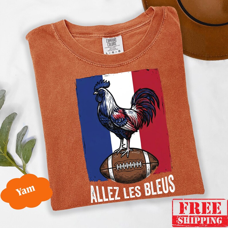 Puede incluir: Una camiseta de manga corta color &oacute;xido con un gr&aacute;fico de un gallo de pie sobre un bal&oacute;n de f&uacute;tbol americano marr&oacute;n sobre un fondo de bandera azul, blanca y roja. El texto "ALLEZ LES BLEUS" est&aacute; impreso en blanco debajo del gr&aacute;fico. Etiqueta Comfort Colors.