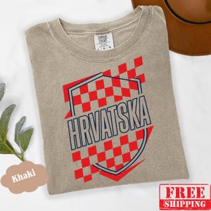 Pu&ograve; includere: Una t-shirt a maniche corte color kaki con un design grafico a scacchi rossi e bianchi all'interno di una forma a scudo con contorno blu scuro. Il testo HRVATSKA &egrave; stampato in lettere maiuscole blu scuro al centro dello scudo.