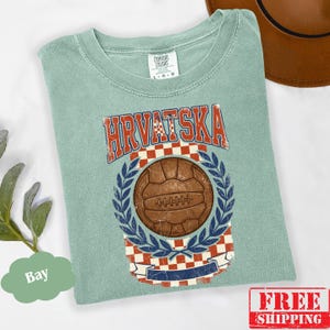 May include: A sage green short-sleeve t-shirt featuring a graphic print with the text HRVATSKA in red letters. The design includes a brown vintage-style soccer ball centered above a red and white checkered pattern, framed by blue laurel leaves.