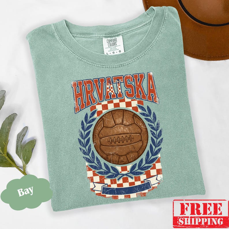 May include: A sage green short-sleeve t-shirt featuring a graphic print with the text HRVATSKA in red letters. The design includes a brown vintage-style soccer ball centered above a red and white checkered pattern, framed by blue laurel leaves.
