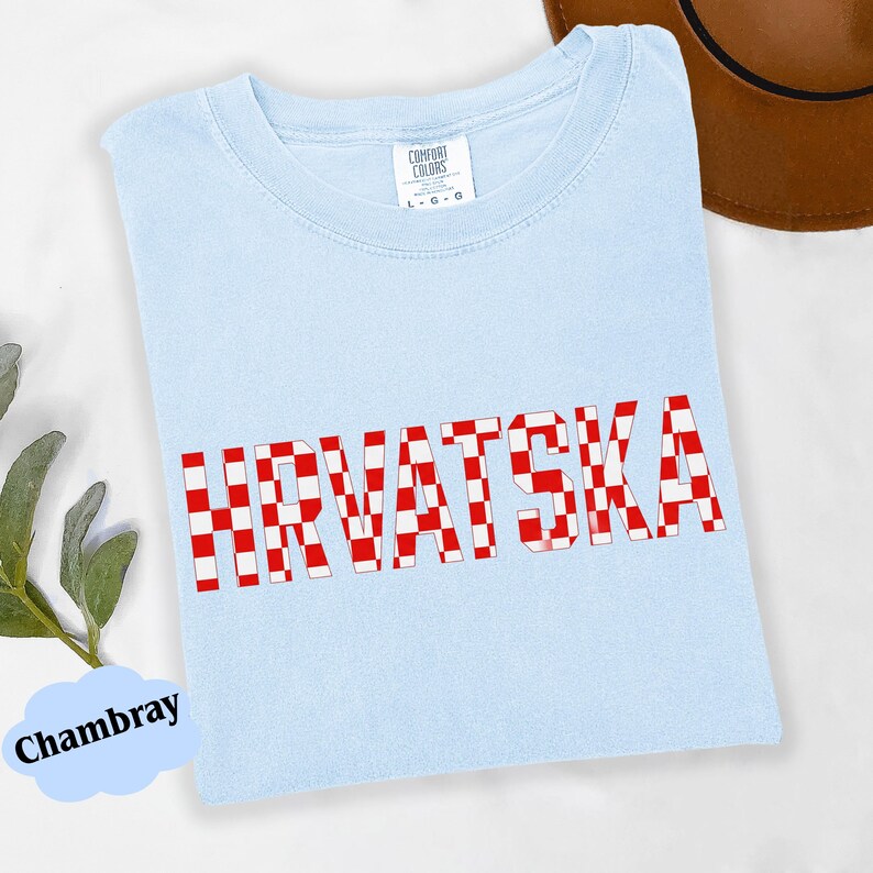 Może przedstawiać: Jasnoniebieska koszulka z napisem "HRVATSKA" w czerwono-białą szachownicę. Koszulka jest złożona i ma metkę z napisem "Comfort Colors". Brązowy kapelusz jest częściowo widoczny w tle.
