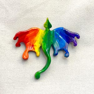 May include: A rainbow-colored dragon-shaped ornament featuring a glossy finish. The creature has a central green body with a long tail, and wings that transition from red and orange on the left to blue and purple on the right side of the figure.