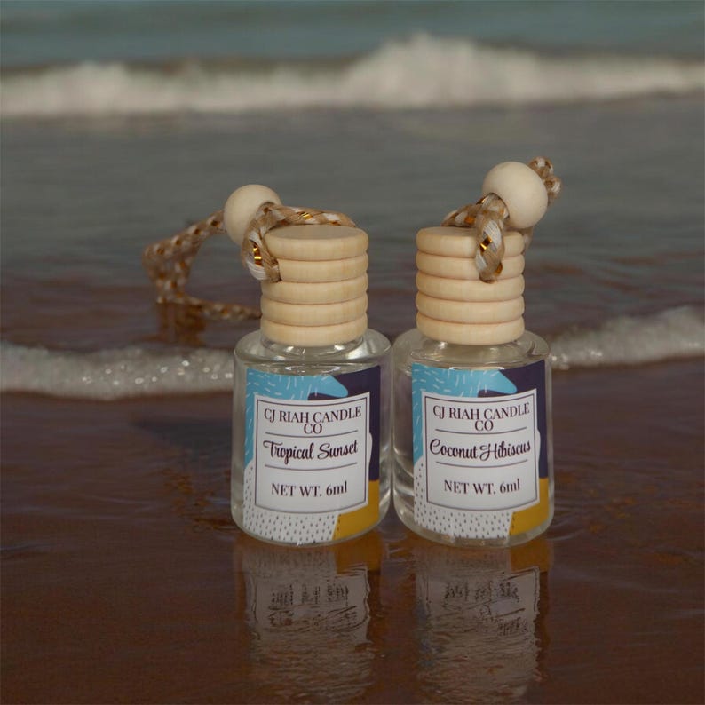 May include: Two small glass bottles of CJ Riah Candle Co. air fresheners, one labelled "Tropical Sunset" and the other "Coconut Hibiscus." Each bottle has a wooden cap and a decorative string. The net weight is 6ml.