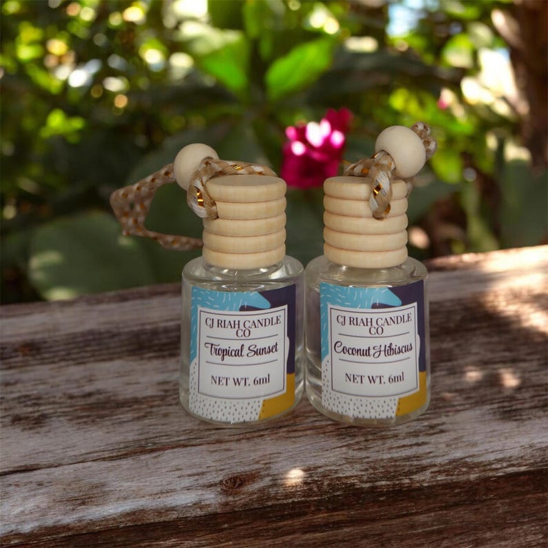 May include: Two small glass bottles with wooden tops and hanging cords. The labels read "CJ Riah Candle Co" and list scents "Tropical Sunset" and "Coconut Hibiscus". Each bottle contains 6ml of product. The background is blurred with green foliage.