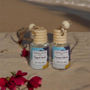 May include: Two small glass bottles with wooden tops and hanging cords. The labels read "CJ Riah Candle Co" and "Tropical Sunset" and "Coconut Hibiscus". Each bottle contains 6ml of product. The bottles are on a sandy surface with red flowers.