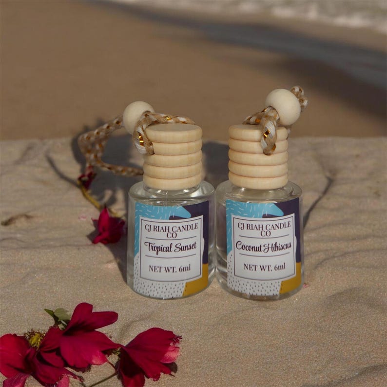 May include: Two small glass bottles with wooden tops and hanging cords. The labels read "CJ Riah Candle Co" and "Tropical Sunset" and "Coconut Hibiscus". Each bottle contains 6ml of product. The bottles are on a sandy surface with red flowers.