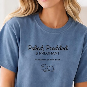 Peut inclure: T-shirt bleu avec l'inscription "Poked, Prodded & Pregnant" et une illustration de bébé. Le t-shirt comprend également le texte "IVF Miracle Coming Soon". Le t-shirt est fait d'une matière douce.
