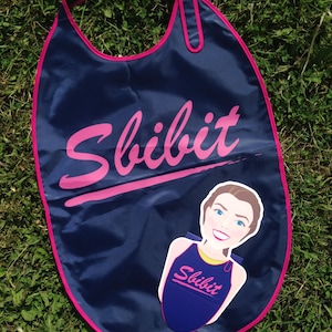 Puede incluir: Una capa azul marino con ribete rosa y la palabra "Sbibit" en escritura rosa. Un recorte de una persona con un atuendo a juego está adjunto. La capa está sobre hierba verde.