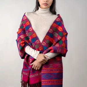 Puede incluir: Un chal vibrante con diseños en tonos fucsia, rojo, naranja, azul y verde. El chal presenta un diseño en forma de hoja en la parte superior y un patrón de rayas con flecos en la parte inferior. La modelo lleva un cuello alto beige.