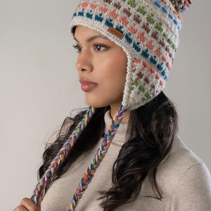 Puede incluir: Un gorro de punto colorido con orejeras y un pompón. El gorro presenta un patrón repetido de cuadrados en varios colores, incluyendo azul, naranja, verde y marrón. Cordones trenzados multicolores se extienden desde las orejeras.
