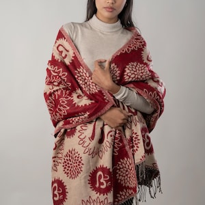 Può includere: Uno scialle bordeaux e beige con un motivo floreale e di lettere. Lo scialle ha un bordo con frange ed è drappeggiato sulle spalle. La combinazione di colori è un mix di bordeaux e beige, creando un'estetica calda e invitante.
