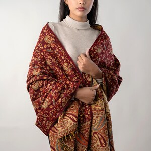 Könnte beinhalten: Ein bordeauxfarbener Schal mit einem aufwendigen Paisley-Muster in Gold-, Orange- und Blautönen. Der Schal hat einen Fransenrand und ist über die Schultern drapiert. Ein warmes und stilvolles Accessoire.