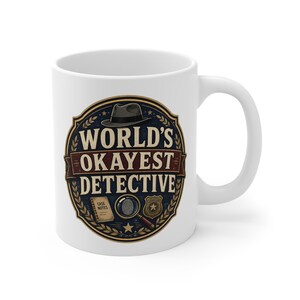 May include: A white ceramic mug featuring a circular graphic with a fedora hat, a magnifying glass, a badge, and a notebook labeled CASE NOTES. The text reads WORLD'S OKAYEST DETECTIVE in bold lettering, surrounded by a decorative laurel leaf border.