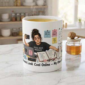 Könnte beinhalten: Ein weißer Keramikbecher mit einer grafischen Illustration eines Social-Media-Managers, der an einem Schreibtisch mit Laptop, Planer und Haftnotizen arbeitet. Der Text lautet: Brands Look Cool Online - My Own Life Is A Mess. Der Becher hat eine glatte Oberfläche.