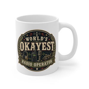 May include: A white ceramic mug featuring a circular graphic with the text "WORLD'S OKAYEST RADIO OPERATOR". The design includes illustrations of headphones, a handheld radio, a vintage microphone, and a telegraph key, all in muted tones.