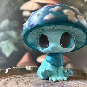May include: A whimsical, hand-painted mushroom figurine in shades of blue and white. The mushroom has large, expressive eyes and a charming, seated pose. The figurine is set against a blurred background of other mushrooms and foliage.