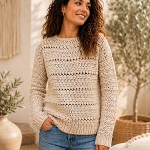 May include: A beige long-sleeved jumper featuring a textured crochet pattern with horizontal rows of openwork stitches and solid bands. The garment has a round neckline and ribbed cuffs, paired with blue denim jeans.